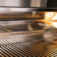 Close-up of TrueFlame 32 inch built-in stainless steel grill interior with cooking grate and warming rack tray illuminated by halogen light